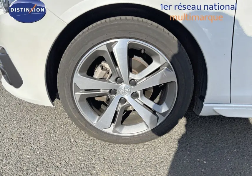 Gros plan sur la roue avant gauche d'une Peugeot 308 blanc nacré, mettant en valeur la jante alliage moderne.