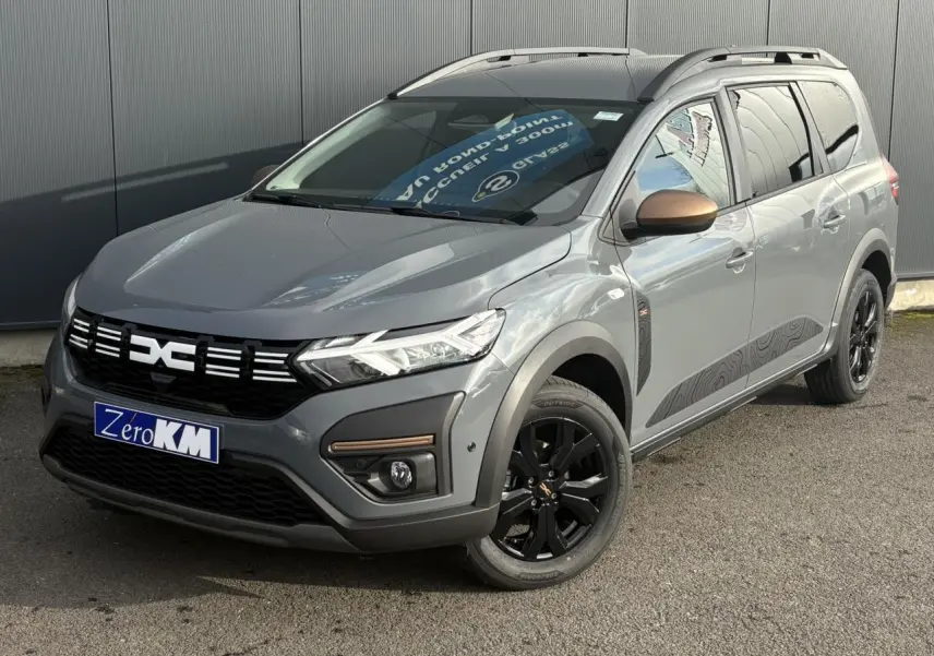 Dacia Jogger Gris Urbain en 3/4 avant droit avec jantes noires et barres de toit modulaires visibles.