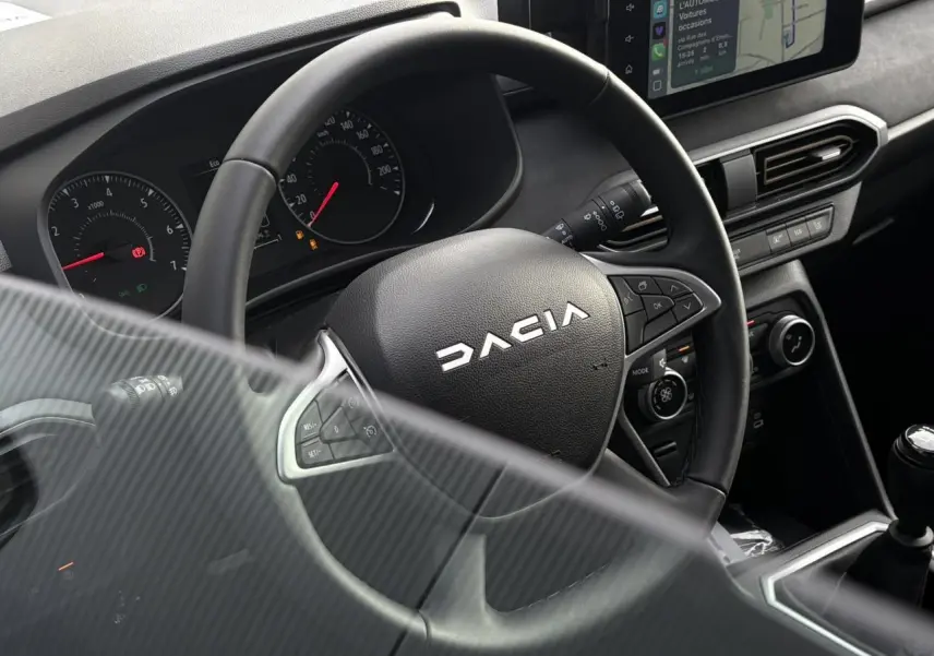 Vue rapprochée du volant DACIA et du tableau de bord avec écran tactile et levier de vitesses manuelle.