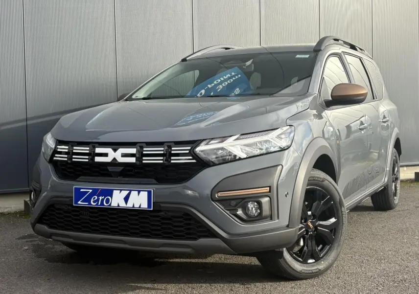 Dacia Jogger Gris Urbain en 3/4 avant droit avec jantes noires et barres de toit modulaires visibles.