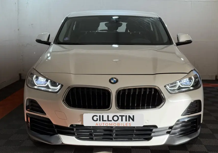 BMW X2 sDrive18i Lounge blanc vue de face avec phares LED allumés dans un garage fermé.