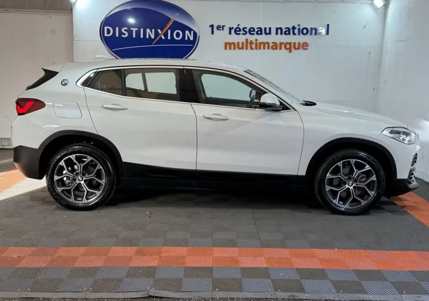 BMW X2 sDrive18i blanc vue de profil côté gauche, avec jantes alliage et feux arrière allumés dans un showroom.