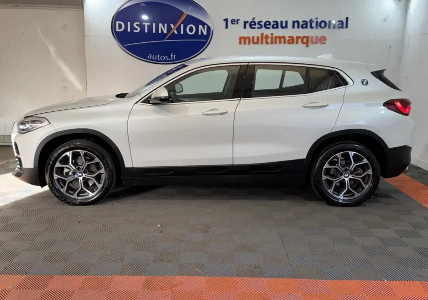 Profil côté gauche d’un BMW X2 sDrive18i Lounge blanc, avec jantes alliage et feux arrière LED visibles en intérieur showroom.