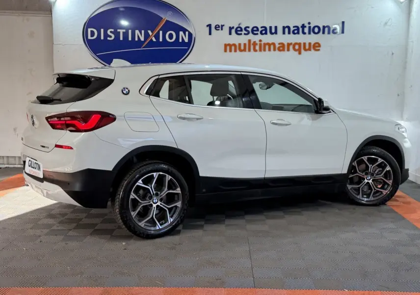 Profil droit d'un BMW X2 sDrive18i Lounge blanc, avec jantes alliage et feux arrière LED visibles en intérieur showroom.