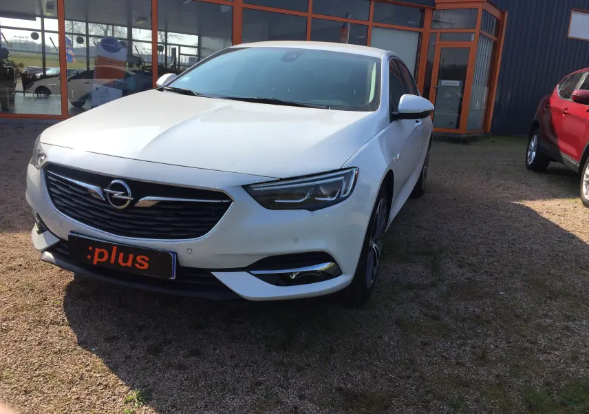 Opel Insignia Grand Sport blanc nacré vue 3/4 avant droit, garée devant un bâtiment avec plaque :plus visible.