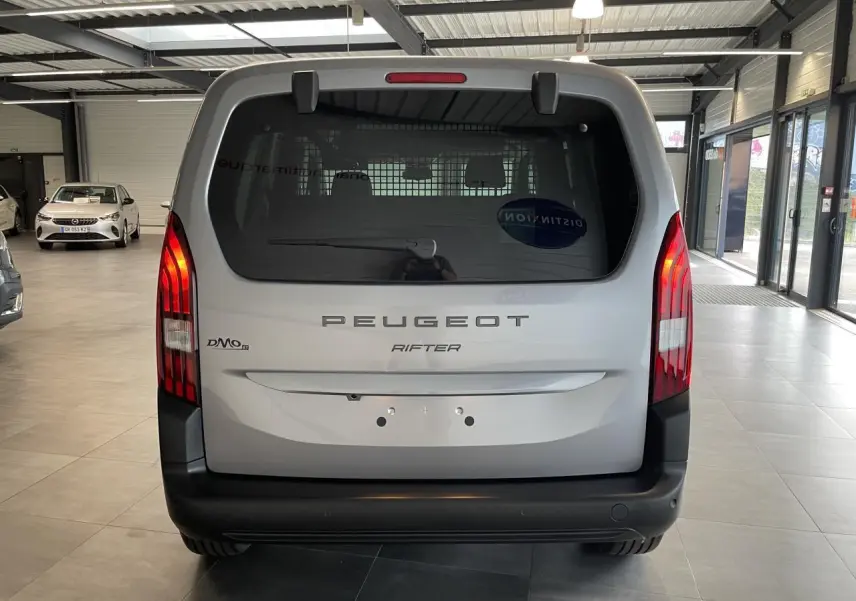 Vue arrière d’un Peugeot Rifter gris Artense avec hayon vitré et feux arrière verticalisés en intérieur showroom.