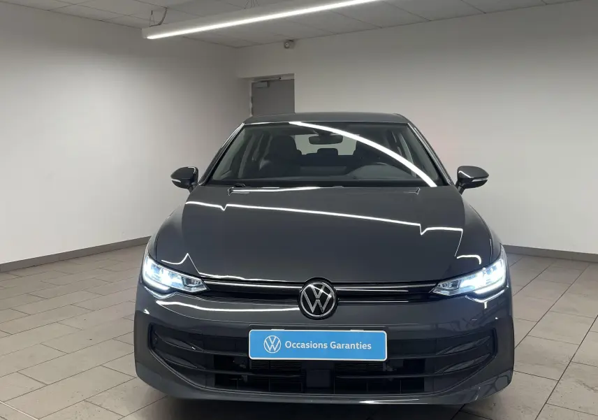 Vue avant d'une Volkswagen Golf 1.5 TSI Gris Dauphin avec phares allumés dans un showroom lumineux.