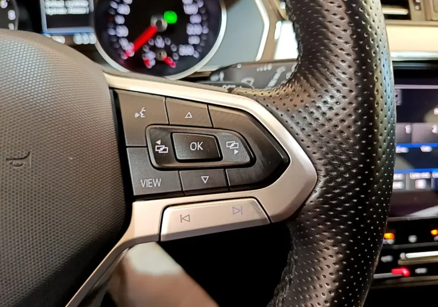 Gros plan sur les commandes gauche du volant en cuir perforé de la Volkswagen Passat hybride blanche, avec tableau de bord digital en arrière-plan.