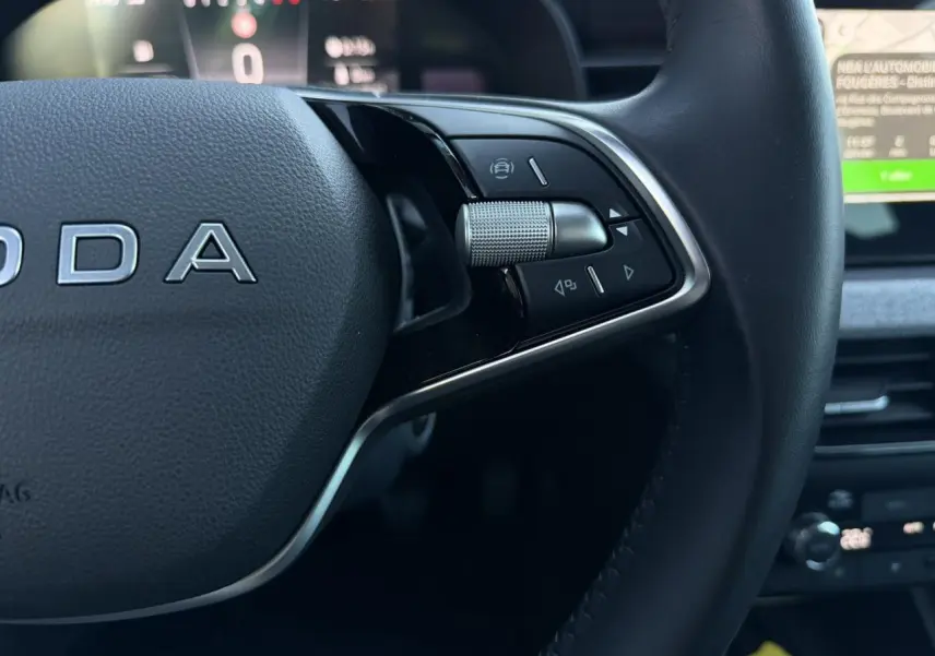 Gros plan sur le volant cuir noir multifonctions de la Skoda Scala TSI 116, avec commandes intégrées et tableau de bord numérique flou en arrière-plan.