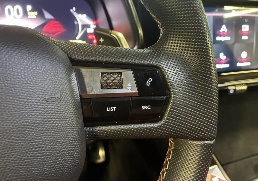 Gros plan sur la partie gauche du volant en cuir perforé noir du DS7 E-TENSE 2024 avec commandes intégrées et tableau de bord digital flou.