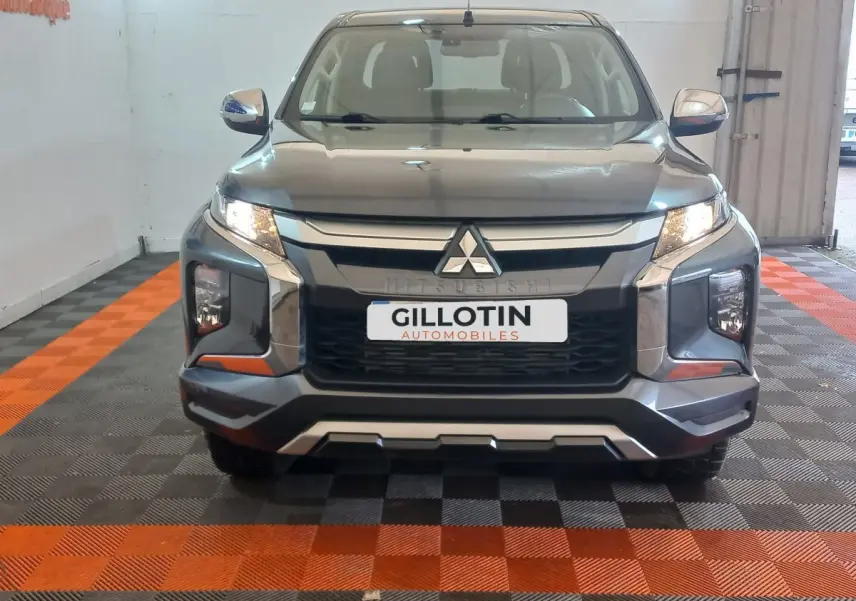 Vue frontale d'un Mitsubishi L200 gris foncé avec calandre chromée et phares allumés en intérieur showroom.