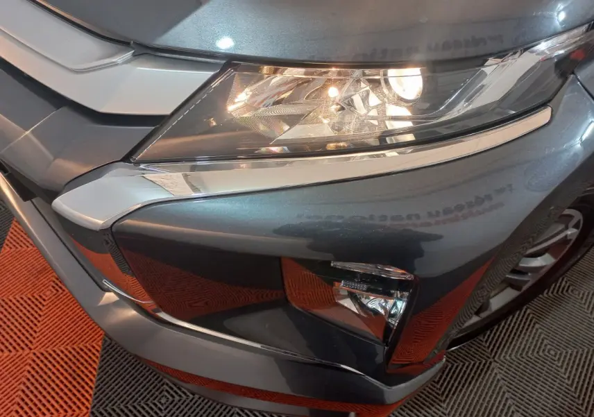 Gros plan sur l'angle avant droit du Mitsubishi L200 gris foncé, mettant en valeur le phare allumé et la calandre chromée.