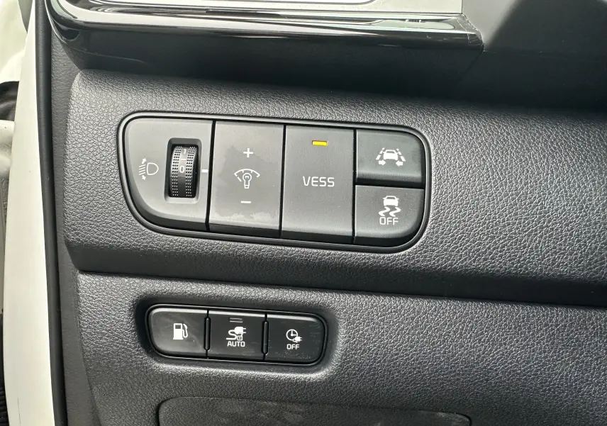 Gros plan sur les commandes noires du tableau de bord du Kia Niro hybride rechargeable blanc céleste, avec boutons d'assistance et réglages lumineux.
