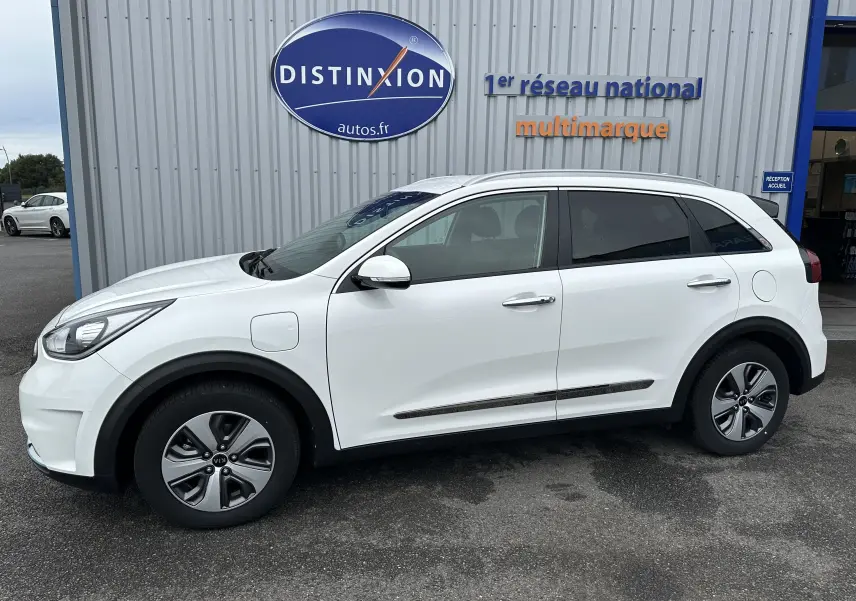 Vue de profil côté gauche d'un KIA Niro blanc céleste hybride rechargeable avec jantes spécifiques et prise de recharge visible.