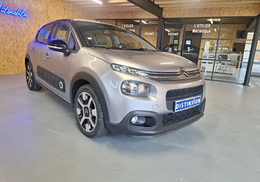 Citroën C3 gris en 3/4 avant droit avec jantes alu et protections latérales noires dans un garage.