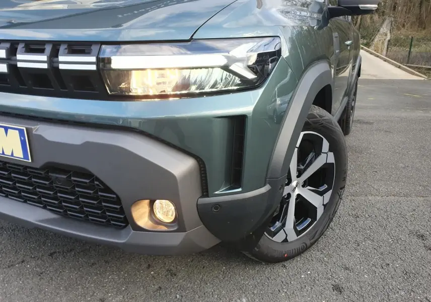 Gros plan sur l'avant droit du Dacia Duster vert cèdre, mettant en valeur le phare allumé et la jante alliage 18 pouces.