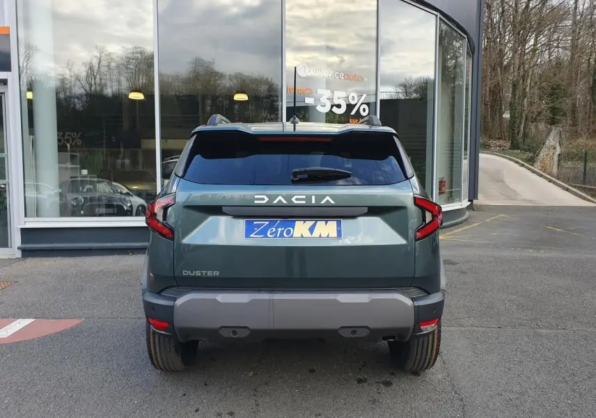 Vue arrière d'un Dacia Duster 2026 vert cèdre avec barres de toit et feux LED distinctifs, stationné devant une vitrine.