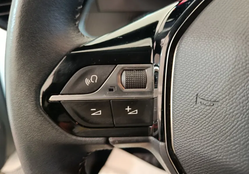 Gros plan sur les commandes gauche du volant cuir noir de la Peugeot 208 blanc, avec boutons de volume et reconnaissance vocale.