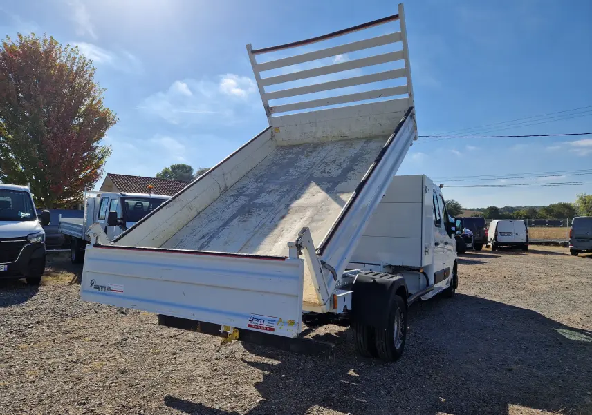 Vue 3/4 arrière droite d’un Renault Master blanc avec benne relevée et coffre, roues jumelées visibles, sur terrain extérieur.