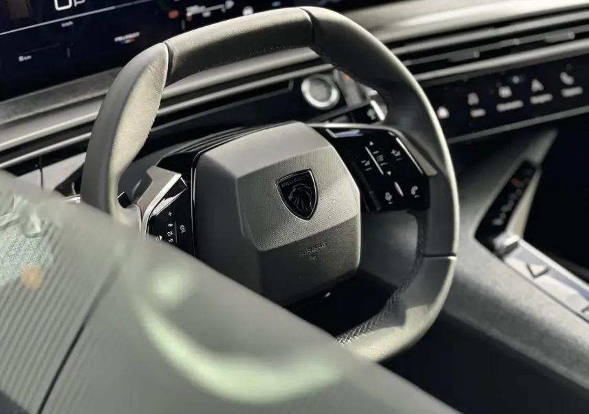 Gros plan sur le volant compact cuir noir avec logo Peugeot du Peugeot 3008 Hybrid 2025, intérieur moderne et écran panoramique visible.
