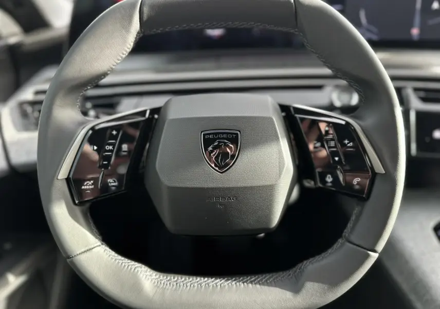 Gros plan sur le volant cuir gris du Peugeot 3008 Hybrid 2025 avec commandes intégrées et logo lion central.