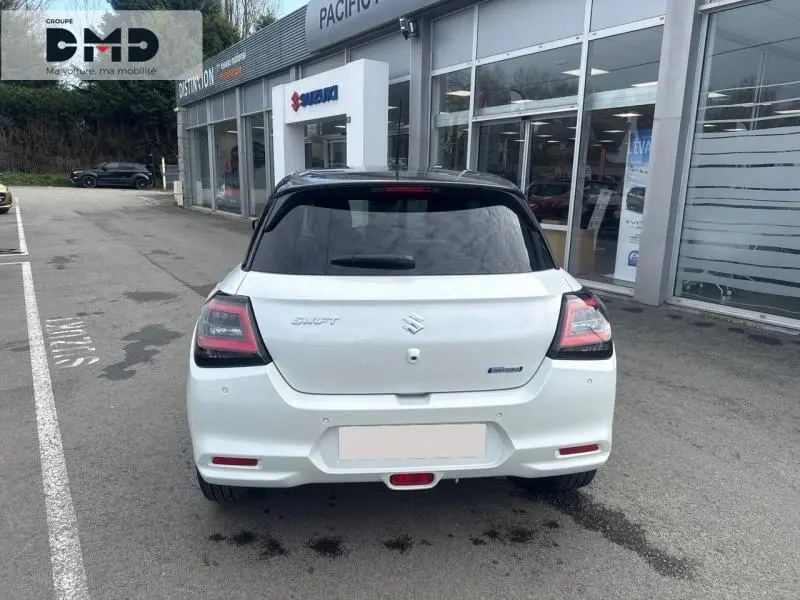 Vue arrière d'une Suzuki Swift blanche à toit gris foncé, stationnée devant une concession Suzuki.
