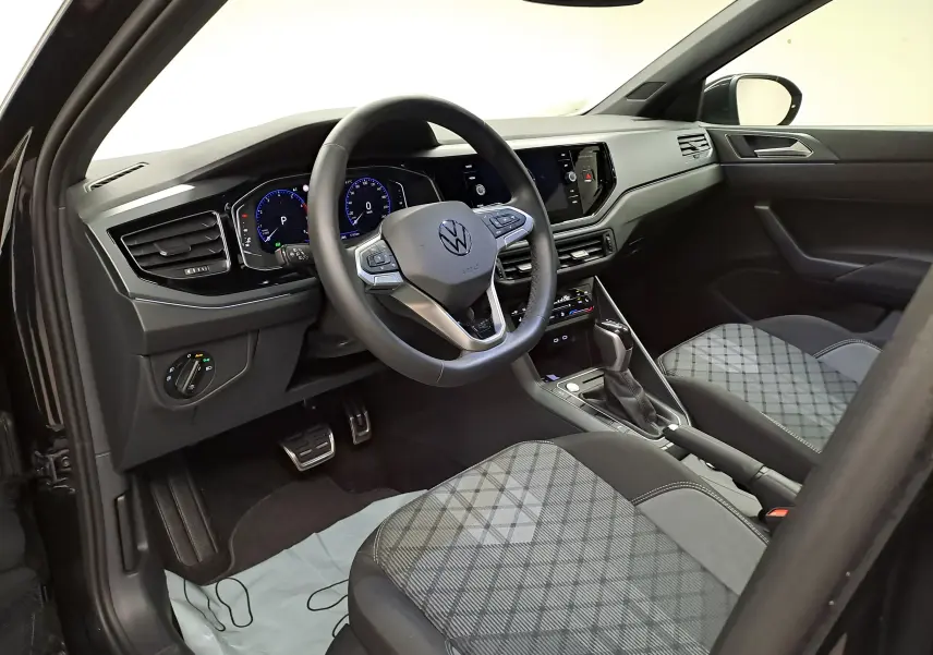 Vue intérieure côté conducteur du Volkswagen Taigo 2025, tableau de bord noir avec sièges tissu à motif gris et volant cuir multifonctions.