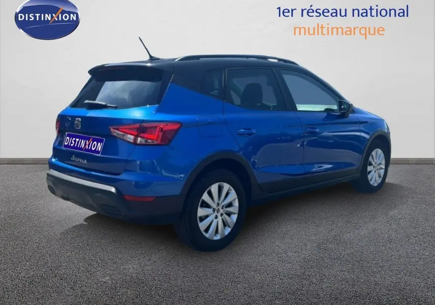 Vue 3/4 arrière droite d'une SEAT Arona bleu saphir métal avec toit noir, stationnée sur sol gris devant un mur blanc.