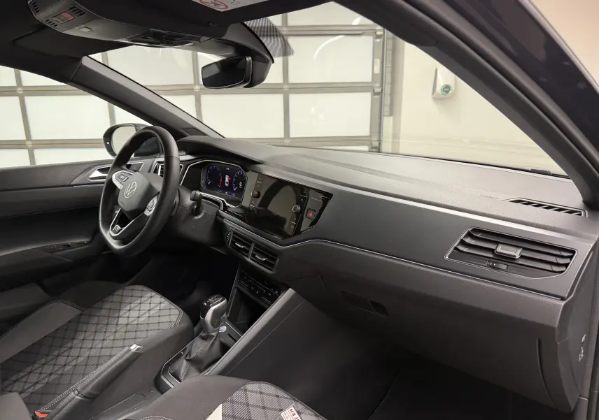 Intérieur de la Volkswagen Taigo 2025 vu côté conducteur, tableau de bord noir avec écran tactile et sièges tissu à motif gris.