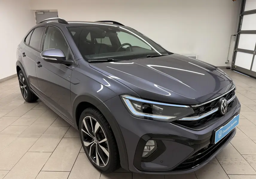 Volkswagen Taigo gris cendre en 3/4 avant droit, avec jantes alliage et calandre R-Line visible en intérieur showroom.