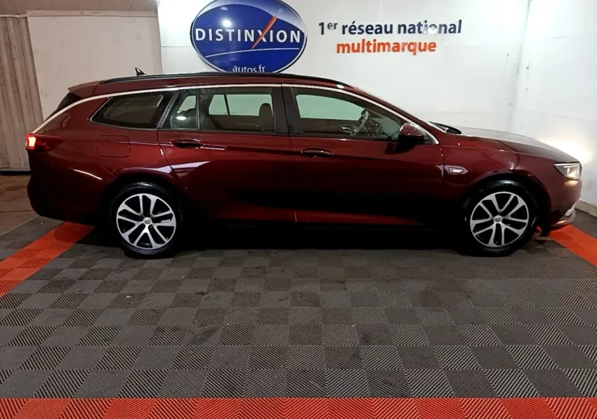 Vue latérale droite d'une Opel Insignia break rouge 2019 dans un showroom avec sol à damier gris et rouge.