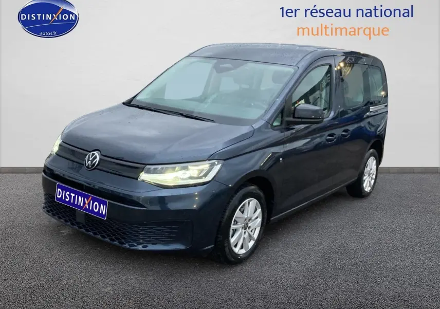 Volkswagen Caddy bleu métallisé vu de 3/4 avant droit, phares allumés sur fond neutre intérieur.