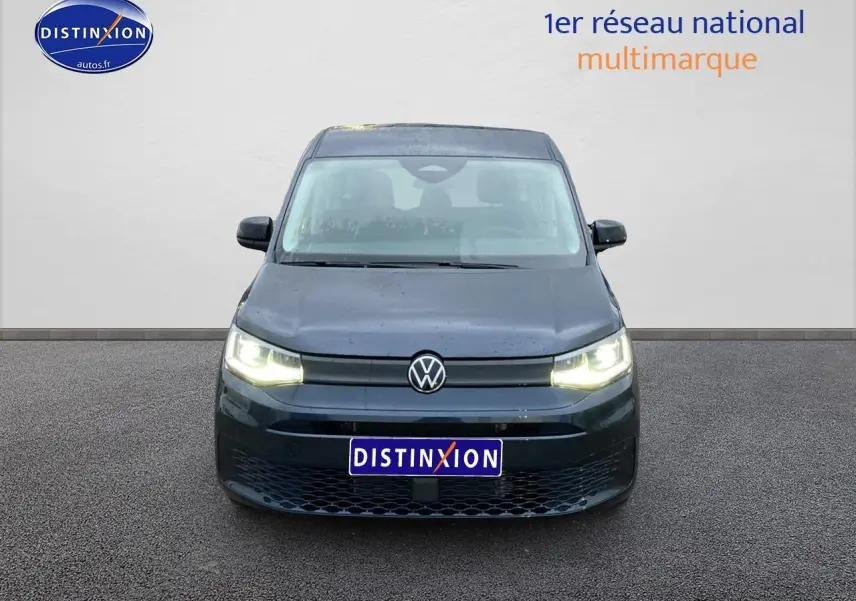 Vue frontale d'un Volkswagen Caddy 2.0 TDI bleu métallisé avec phares allumés sur fond neutre.
