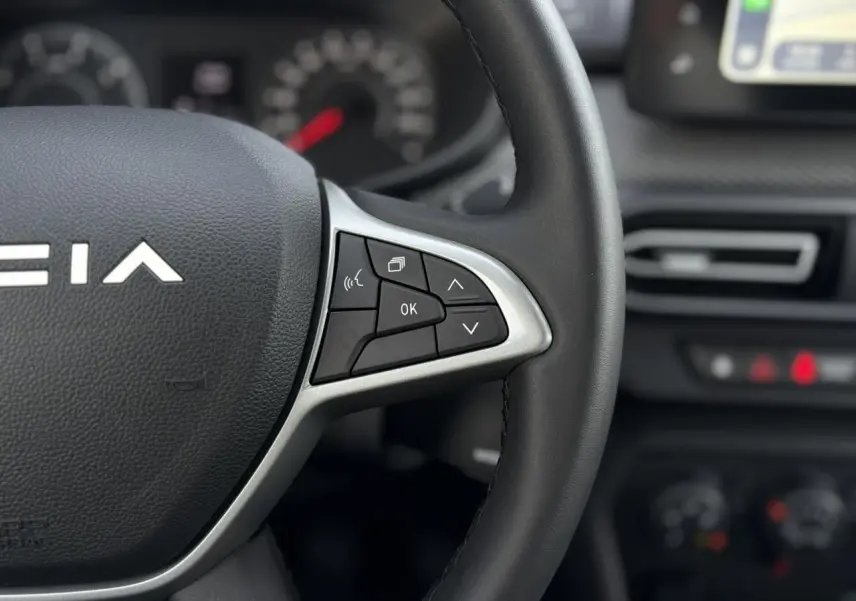Gros plan sur le volant noir du Dacia Jogger 2024 avec commandes multifonctions et tableau de bord flou en arrière-plan.