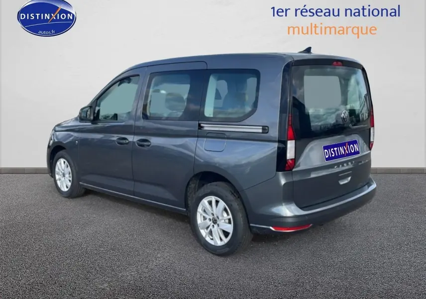 Volkswagen Caddy gris indien vu en 3/4 arrière droit avec vitres teintées et barres de toit visibles.