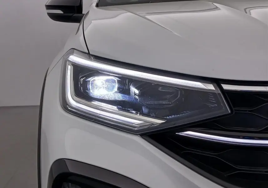 Gros plan sur le phare avant gauche allumé d'un Volkswagen Taigo blanc pur avec toit noir et calandre noire.