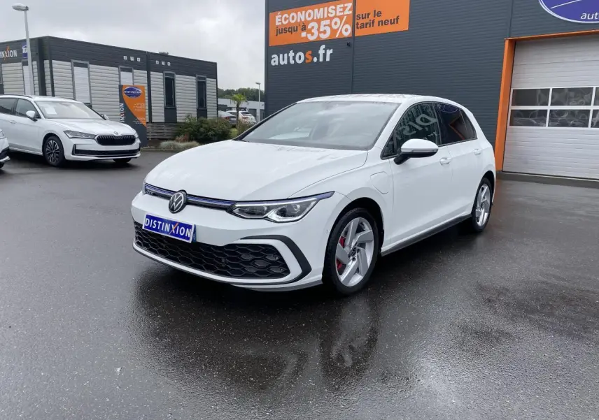 Volkswagen Golf 1.4 eHybrid blanche vue en 3/4 avant droit sur parking humide devant un bâtiment commercial.