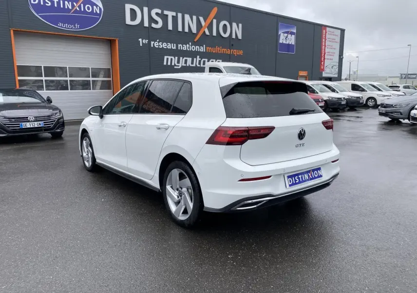 Vue 3/4 arrière droite d'une Volkswagen Golf VIII GTE blanche pur avec vitres surteintées sur parking humide.