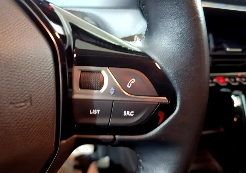 Gros plan sur les commandes gauche du volant noir de la Peugeot 208 1.5 BlueHDi 100 S&S Premium, avec boutons d’appel et réglages audio.