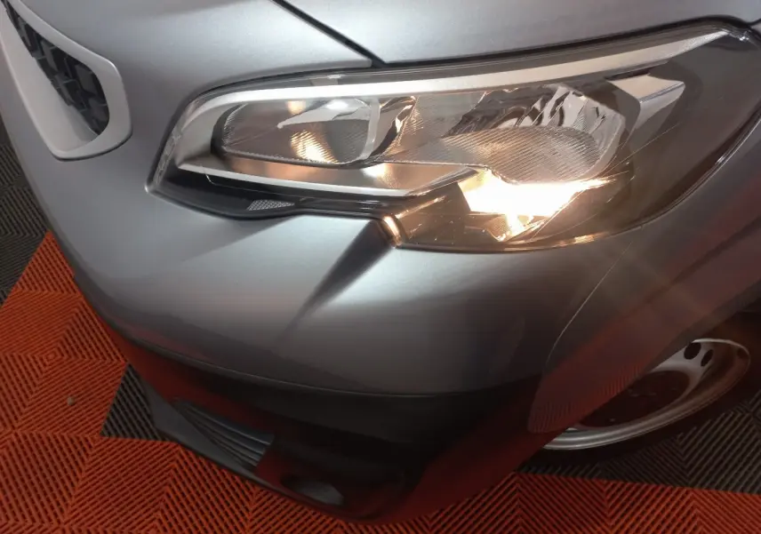 Gros plan sur le phare avant droit du Peugeot Traveller gris clair, avec partie de la calandre et roue visible.