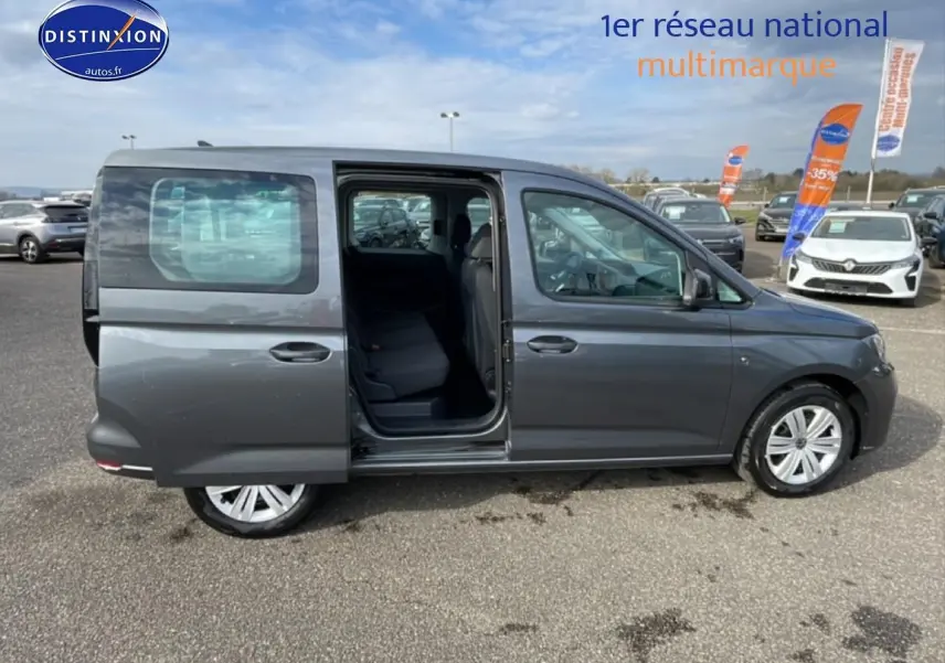 Profil droit d’un Volkswagen Caddy gris indien 2025 avec porte coulissante arrière ouverte sur parking.