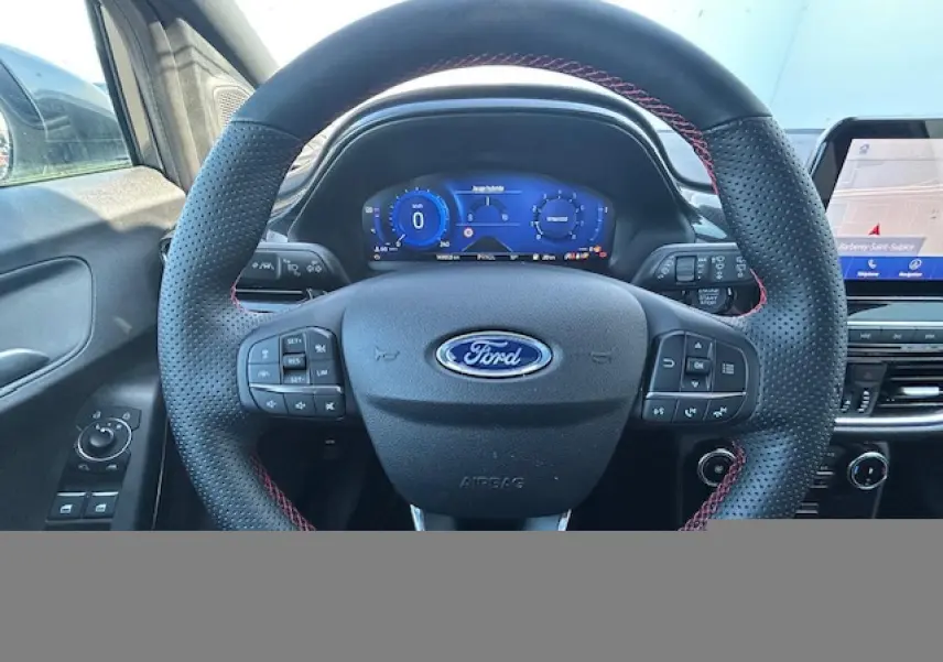 Vue intérieure centrée sur le volant perforé noir avec surpiqûres rouges du Ford Puma gris, tableau de bord numérique et écran tactile.