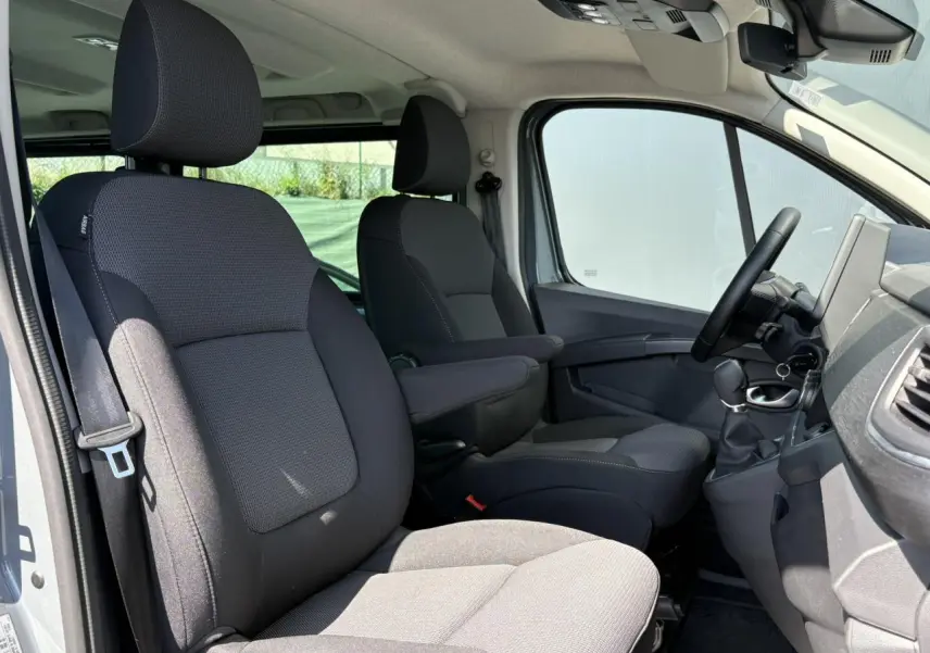 Vue intérieure côté conducteur du Renault Trafic Fourgon 2024, sièges confort gris foncé avec accoudoirs et volant visible.