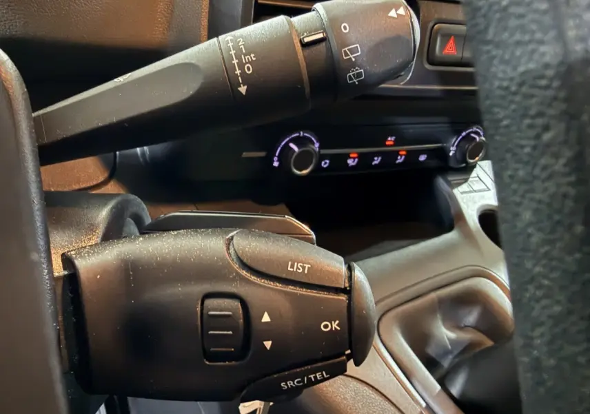 Gros plan sur les commandes au volant noir du Peugeot Rifter blanc, avec tableau de bord et commandes climatisation en arrière-plan.