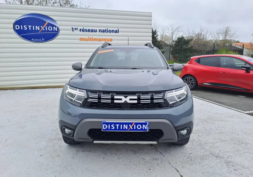Vue avant d'un Dacia Duster 2024 gris urbain avec calandre noire et phares LED allumés sur parking.