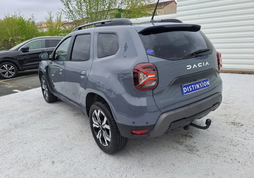 Vue 3/4 arrière droite d'un Dacia Duster gris urbain 2024 avec barres de toit et attelage visible.