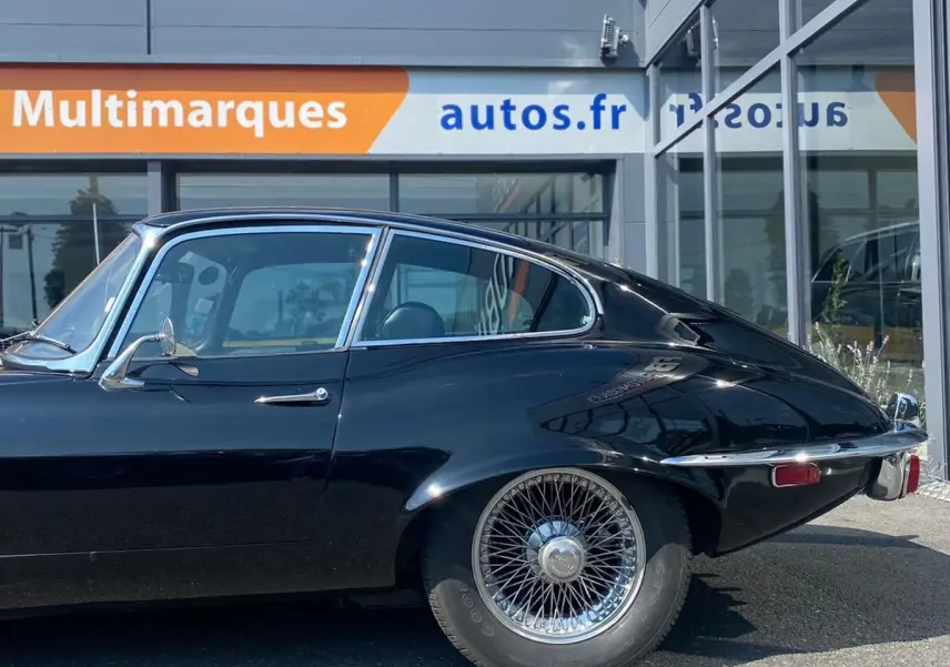 Profil côté gauche d'une Jaguar S-Type Type E Coupé noir de 1970 avec jantes à rayons chromés, garée devant un bâtiment moderne.
