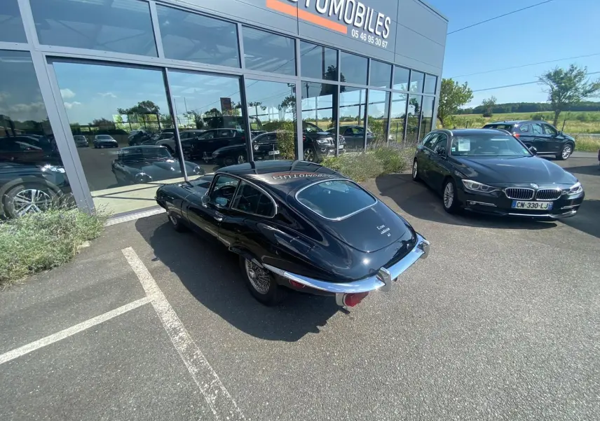 Jaguar Type E Coupé noir vue 3/4 arrière droit, garée devant un showroom avec jantes à rayons chromés.