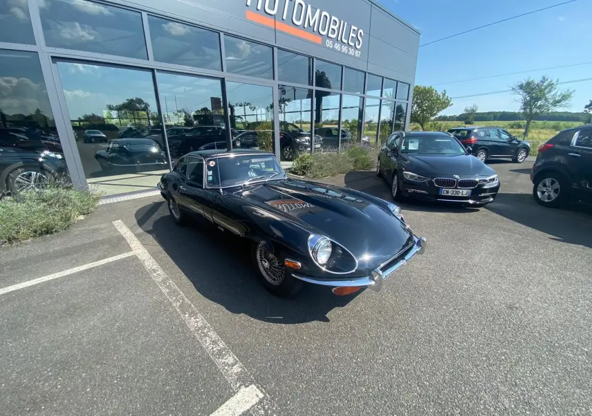 Jaguar Type E Coupé 4.2 Série 2 noire vue en 3/4 avant droit sur parking devant un showroom automobile.