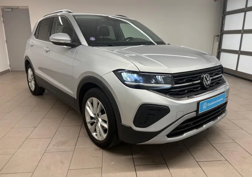 Volkswagen T-Cross 2024 argent reflet, vue 3/4 avant droit en intérieur avec phares LED allumés.